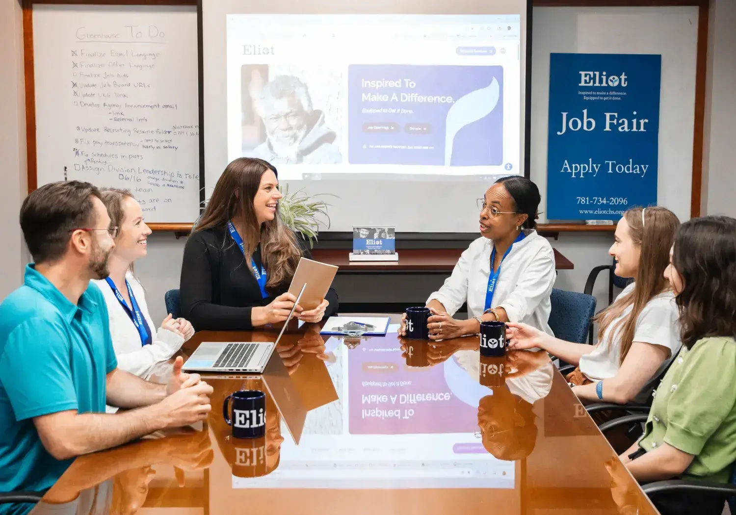 A diverse group of six people are seated around a polished wooden conference table, engaged in a discussion. A laptop, more "Eliot" mugs, and papers are on the table. Behind them, a large screen displays a website or presentation with the word "Eliot" and "Inspired To Make A Difference." To the left of the screen, a whiteboard lists "Crossover To-Do" items. On the far right, a blue poster advertises an "Eliot Job Fair."
